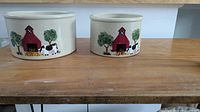 Two ceramic crocks with farm scene including red barn, cows, and trees on wooden table