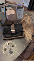 Photo showing a black leather handbag with silver buckle clasp, a blonde wavy Remy real hair ponytail, and two skincare product boxes on a wooden table.