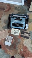 Smith Corona light blue typewriter placed on colorful carpet, accompanied by four original brochures laid out alongside the case bottom half.