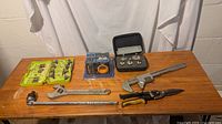 Full table of assorted hand tools including wrenches, pliers, socket wrench extension, screwdriver bits in sealed pack, and pipe fitting set in black case.