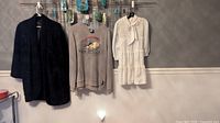 Three vintage clothing items hanging on a wall: black beaded coat, grey Astro Boy sweatshirt, white smock dress