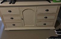 Front view of cream-colored wooden dresser showing 8 drawers and central cabinet door with black metal handles