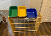 Yellow children's toy rack with three plastic bins (green, yellow, blue) placed on top against a white door.
