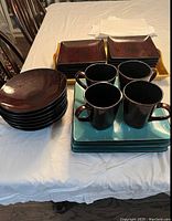 Brown round and square ceramic plates stacked with four black mugs on blue coasters on a white tablecloth