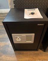 Front view of matte black Stalwart electronic safe with silver keypad and handle. User manual is placed on top of the safe.
