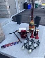 Home decor items arranged on concrete, including black square planters, a multicolored tall vase, red glass candle holders, a red glass vase, two black lantern-style lights and a small metallic decorative box along with a scooter visible partially.