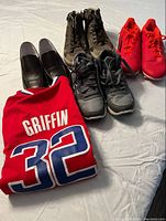Four pairs of kids shoes and a red Griffin 32 jersey laid out side by side on a flat surface.