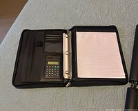 Opened black leather binder revealing interior featuring a calculator, several card slots, ID window, pen holder, and a metal 3-ring binder holding lined notepad sheets.