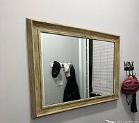 Wall-mounted rectangular mirror with a wide light wood frame, reflecting a coat rack and window blinds.