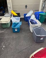 Wide view of the assortment of plastic storage totes and lids on driveway, showing variety of colors and sizes.