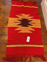 Full view of the red woven mat with central yellow and dark brown geometric pattern, fringed ends, laid flat on wood floor showing entire piece with seller tag