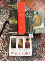 Four art books placed on a patterned rug: Paintings of the Louvre with a classical painting cover, Jansons History of Art in red spine, Wyeth art book in softcover at bottom, and decorative cover box.