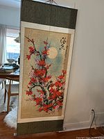 Full view of the Asian scroll showing the floral design and porcelain roller with green cloth border, hanging indoors.