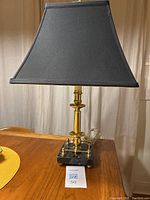 Front view of the brass candlestick lamp on the black marble base with black linen shade, placed on a wooden surface, with the lamp tag showing lot number.