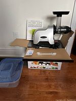 Omega juicer set up on cardboard with plastic container and cleaning tool in front, book in background.