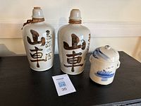 Two vintage Japanese saki bottles with brown calligraphy and rope around necks, alongside a small Chinese blue and white tea caddy with lid, all on a black surface with plain white wall background.