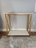 Full front photo of console table with white wood surfaces and gold metal frame resting on a rug against a wall.