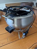 Metallic silver electric fondue pot with attached power cord and multiple forks resting on top, placed on wooden surface.