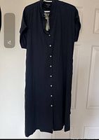 Full view of the navy blue Frank and Oak dress hanging from a door, buttoned up with white buttons down the front, showing the straight cut and long sleeves.