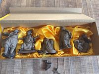 Five dark ceramic Japanese figurines displayed in a box with yellow silk-like lining; one figurine is broken into two pieces lying separately in front of the box.