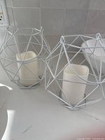Two white metal geometric cage candle holders with flameless LED candles inside, sitting on a white surface.