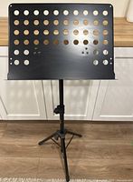 Front view of the black metal music stand with perforated sheet holder and adjustable tripod base.
