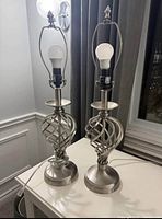 Two metal table lamps with spiral cage bases, bulb installed but no shade, placed on a white surface near window and wall.