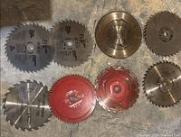 Nine assorted circular saw blades including silver and red blades with various tooth designs, shown on a wooden surface.