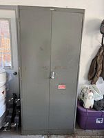 Closed grey steel cabinet with double doors and latch closing mechanism, showing minor wear and some scuff marks on the exterior.
