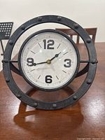 Front view of the steel table clock showing the white dial with black numerals and black hour and minute hands, secured within a riveted circular steel frame.