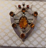 Front view shows heart-shaped silver brooch with claw-set oval citrine center, six round citrines around, and crown with small red and white stones on top.