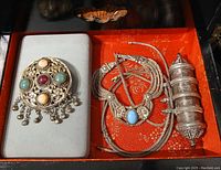 Group shot of antique tribal silver jewelry in a red lined box showing round brooch with stones, multiple necklaces with woven chains and cylindrical pendant.