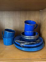 Photo of stacked blue enamel mugs and plates with cutlery on top showing the lot contents and style