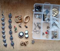 Top-down view of the assortment including two large bracelets, several brooches, earrings, rings, and a plastic organizer with small loose jewelry pieces.