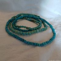 Full view of both coiled blue-green stone bead necklaces on white cloth background.