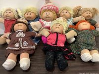 Group photo showing eight 1980s Cabbage Patch Kids dolls lying down. Dolls have various yarn hair colors and outfits in vintage style suggesting signs of moderate to heavy use.