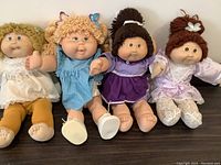 Four Cabbage Patch Kids dolls sitting side by side on a table showing front view with clothing and hair styles visible.