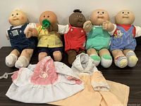 Five preemie Cabbage Patch dolls lined up wearing different outfits with two preemie gowns laid front center.