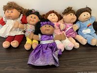 Six Cabbage Patch Kids dolls seated side by side, showing different outfits and hairstyles.