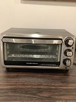 Front view of stainless steel Hamilton Beach toaster oven with closed glass door and control knobs on right side, showing minor discoloration on top surface.