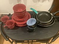 Set of red ceramic dishes, cups, and four non-stick pans on a black shelving unit