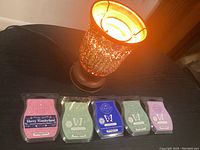 Photo of lit golden glittery Scentsy wax warmer on a black surface with five unopened wax bars lined in front showing front label designs and scents.