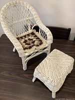 Full view of white wicker doll chair with crocheted cushion and matching wicker stool on dark surface.