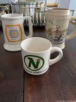 Three white and beige ceramic mugs with handles: Molson Golden Ale logo mug, Minnesota North Stars sports logo mug, and a wildlife scene mug.