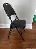 Side view of one black folding chair with cushioned seat and backrest on wood floor against white wall.
