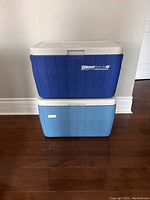Two Coleman coolers stacked vertically: top is dark blue PolyLite 48 model, bottom is light blue vintage cooler, both have white lids.