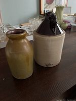 Two ceramic crocks placed side by side on a wooden table. One brown crock with narrow mouth, other white with dark brown top.