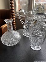 Photo showing three crystal glass pieces placed on a table near a window: a decanter, a pitcher, and a vase with detailed cut patterns.