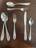 Individual cutlery pieces on wood table showing teaspoons, forks, knives, and serving spoons, demonstrating item types and condition.