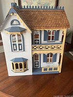 Front view of the wooden dollhouse showing exterior design with windows, blue and beige trim, and shingled roof.